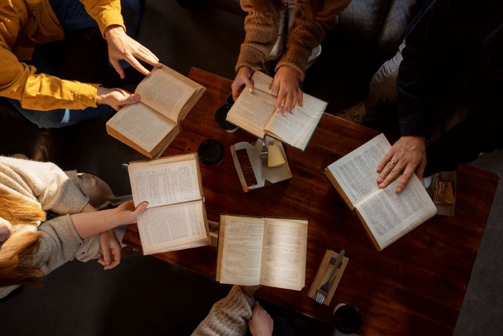 reunión de cinco personas a las cuales se les ve únicamente los brazos y las manos sosteniendo un libro cada una. Están sentadas en una mesa de confitería, el ángulo es desde arriba.